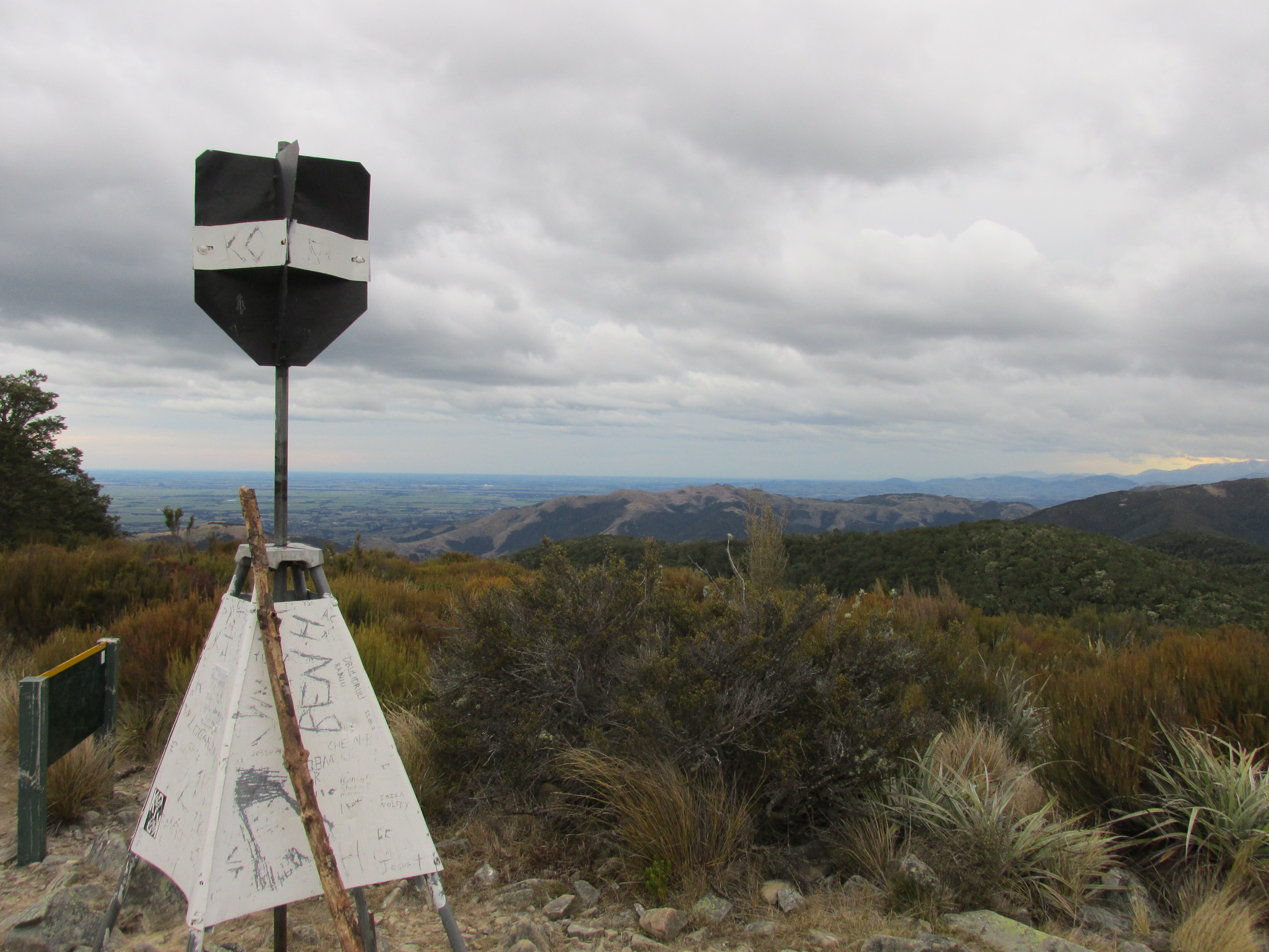 Small picture of a pyrimidal marker object, on the summit of a mountain. A roughly walking-stick shaped stick leans on it.